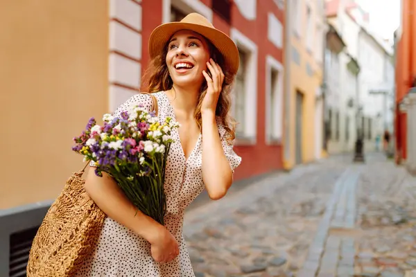 Güneşli Avrupa caddesinde yürüyen şapkalı ve buketi olan güzel bir kadın. Genç turist dışarıda eğleniyor ve eğleniyor. Yürüyen konsept, seyahat.