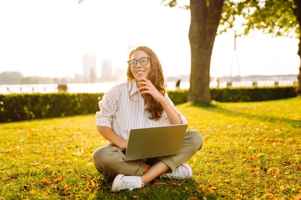 Laptoplu genç bir kadın yeşil çimlerle uğraşıyor. Neşeli kadın serbest yazar gün batımında dışarıda çalışıyor. Eğitim, iş, serbest çalışma kavramı.