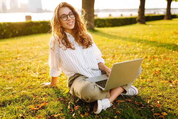 Laptoplu genç bir kadın yeşil çimlerle uğraşıyor. Neşeli kadın serbest yazar gün batımında dışarıda çalışıyor. Eğitim, iş, serbest çalışma kavramı.