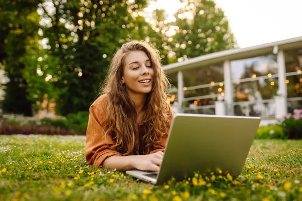 Bilgisayarlı genç bir kadın parkta yeşil çimlerde çalışıyor. Dizüstü bilgisayarı olan serbest çalışan bir kadın parkta güneşli havanın tadını çıkarıyor. Uzaktan çalışma konsepti, serbest yazar. Yaşam biçimi.