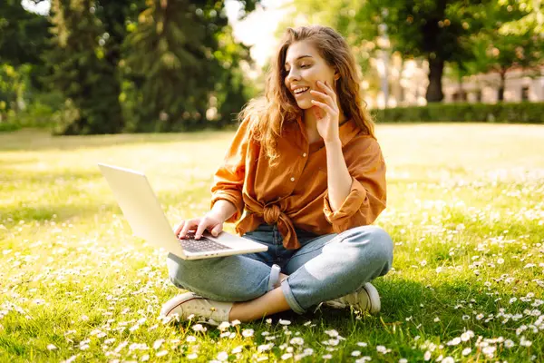 Bilgisayarlı genç bir kadın parkta yeşil çimlerde çalışıyor. Dizüstü bilgisayarı olan serbest çalışan bir kadın parkta güneşli havanın tadını çıkarıyor. Uzaktan çalışma konsepti, serbest yazar. Yaşam biçimi.