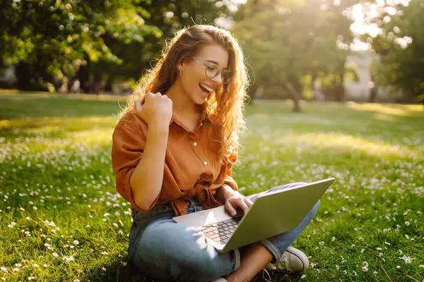 Bilgisayarlı genç bir kadın parkta yeşil çimlerde çalışıyor. Dizüstü bilgisayarı olan serbest çalışan bir kadın parkta güneşli havanın tadını çıkarıyor. Uzaktan çalışma konsepti, serbest yazar. Yaşam biçimi.