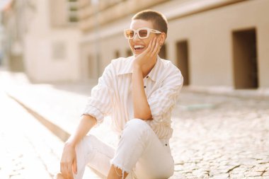 Kısa saçlı şık bir kadın şehrin güneşli sokaklarında poz veriyor. Genç bir turist dışarıda havanın tadını çıkarıyor. Turizm ve moda konsepti.