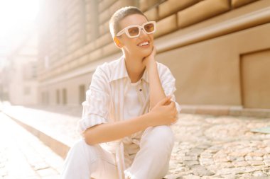 Kısa saçlı şık bir kadın şehrin güneşli sokaklarında poz veriyor. Genç bir turist dışarıda havanın tadını çıkarıyor. Turizm ve moda konsepti.