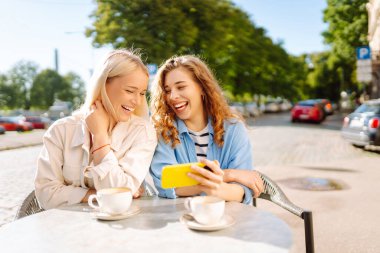 İki güzel kadın selfie çeker ve bir yaz terasında otururken telefonlarında blog çekerler. Genç kız arkadaşlar kahve içer ve güneşli bir günde açık bir kafedeki masada eğlenirler..
