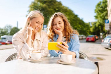 İki güzel kadın selfie çeker ve bir yaz terasında otururken telefonlarında blog çekerler. Genç kız arkadaşlar kahve içer ve güneşli bir günde açık bir kafedeki masada eğlenirler..