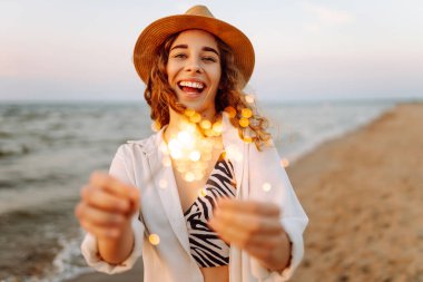Şapkalı mutlu kadın elinde maytaplarla sahilde günbatımının tadını çıkarıyor. Genç bir kadın dışarıda maytaplarla eğleniyor. Tatil ve eğlence konsepti.