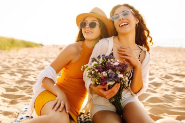 Genç bayan arkadaşlar kumlu sahilde bir buket kır çiçeğiyle eğleniyorlar. İki mutlu kadın dışarıda gün batımını seyrediyor. Tatil, haftasonu ve arkadaşlık konsepti.