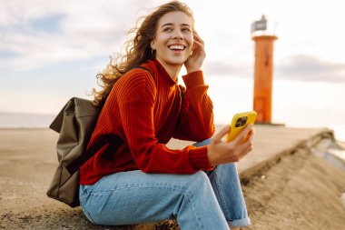 Sırt çantası ve telefonu olan genç bir kadın deniz fenerine karşı kendini özgür hissediyor. Gülümseyen turist selfie çekiyor, günbatımında deniz manzaralı blog yazıyor. Özgürlük kavramı.