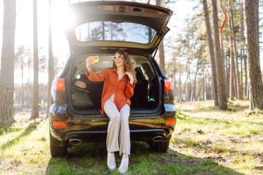 Güneş gözlüklü şık bir kadın bir arabanın bagajında otururken telefonuyla selfie çekiyor. Genç bayan gezgin telefonuyla arabasının yanındaki ormanda günbatımının tadını çıkarıyor. Araba yolculuğu kavramı.