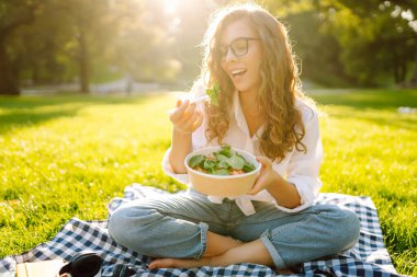 Gözlüklü mutlu kadın parkta bir paspasa oturur ve yeşil salata yer. Genç bir kadın gün batımında parkta atıştırmaktan hoşlanır. Sağlıklı beslenme, rahatlama konsepti.