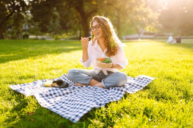 Gözlüklü mutlu kadın parkta bir paspasa oturur ve yeşil salata yer. Genç bir kadın gün batımında parkta atıştırmaktan hoşlanır. Sağlıklı beslenme, rahatlama konsepti.