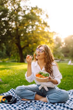 Gözlüklü mutlu kadın parkta bir paspasa oturur ve yeşil salata yer. Genç bir kadın gün batımında parkta atıştırmaktan hoşlanır. Sağlıklı beslenme, rahatlama konsepti.