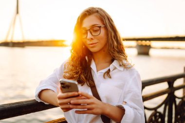 Kıvırcık saçlı, telefon ve gözlüklü bir kadın sette günbatımının tadını çıkarıyor. Telefonu olan güzel bir kadın dışarıda poz veriyor. Yürüme, teknoloji konsepti.