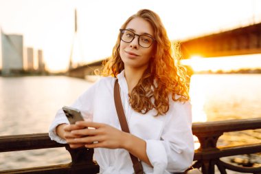 Kıvırcık saçlı, telefon ve gözlüklü bir kadın sette günbatımının tadını çıkarıyor. Telefonu olan güzel bir kadın dışarıda poz veriyor. Yürüme, teknoloji konsepti.