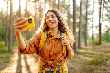 Telefonlu genç bir kadın güneşli bir ormanda selfie çekiyor. Sırt çantalı ve telefonlu bir kadın yürüyüşçü yürüyüş sırasında doğanın tadını çıkarıyor. Günlük konsepti.