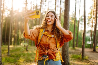 Telefonlu genç bir kadın güneşli bir ormanda selfie çekiyor. Sırt çantalı ve telefonlu bir kadın yürüyüşçü yürüyüş sırasında doğanın tadını çıkarıyor. Günlük konsepti.