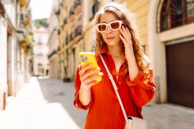 Gözlüklü gülümseyen bir kadın ve Avrupa 'nın güneşli caddelerinde yürüyen bir telefonla. Genç bir turist dışarıda yürümekten zevk alıyor. Turizm konsepti. Yaşam biçimi.