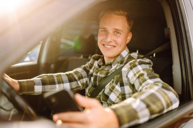 Telefonlu genç adam arabadaki anın tadını çıkarıyor. Neşeli bir adam araba kullanıyor güneşli bir otoparkta telefonla konuşuyor. Ulaşım, teknoloji kavramı.