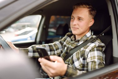 Telefonlu genç adam arabadaki anın tadını çıkarıyor. Neşeli bir adam araba kullanıyor güneşli bir otoparkta telefonla konuşuyor. Ulaşım, teknoloji kavramı.