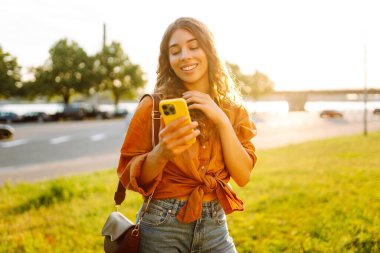 Günbatımında modern giysileri ve telefonu olan mutlu kadın portresi. Güzel bir kadın sohbet ediyor ve dışarıda selfie çekiyor..