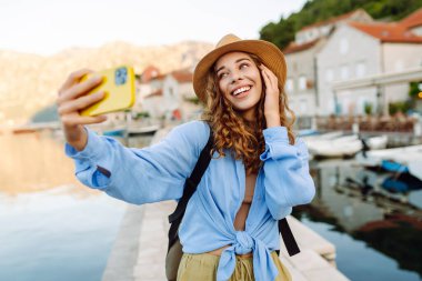 Mutlu kadın poz veriyor ve telefonuyla güneşli bir sette eğleniyor. Telefonlu genç bir kadın dışarıda selfie çekerek şehirde dolaşıyor. Turizm konsepti.