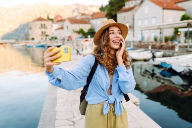 Mutlu kadın poz veriyor ve telefonuyla güneşli bir sette eğleniyor. Telefonlu genç bir kadın dışarıda selfie çekerek şehirde dolaşıyor. Turizm konsepti.