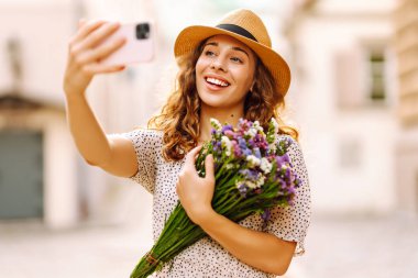 Neşeli kadın güneşli bir günün tadını çıkarıyor ve telefonuyla selfie çekiyor. Bir buket kır çiçeğiyle güzel bir turist güneşli bir şehirde yürüyor. Yaşam biçimi.