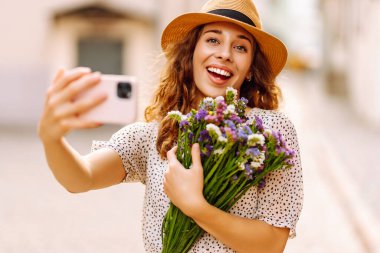 Neşeli kadın güneşli bir günün tadını çıkarıyor ve telefonuyla selfie çekiyor. Bir buket kır çiçeğiyle güzel bir turist güneşli bir şehirde yürüyor. Yaşam biçimi.