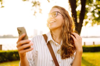 Güneşli bir tepede telefonu olan mutlu bir kadının portresi. Güzel bir kadın dışarıda eğleniyor ve blog yazıyor. Bloglama ve yürüme konsepti.