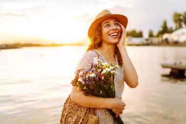 Genç bir kadın gün batımında bir buket yabani çiçekle gülüyor. Çiçek turisti bir buket çiçekle deniz manzarasının tadını çıkarıyor. Keyif ve rahatlama konsepti.