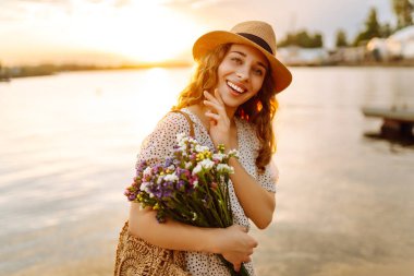 Genç bir kadın gün batımında bir buket yabani çiçekle gülüyor. Çiçek turisti bir buket çiçekle deniz manzarasının tadını çıkarıyor. Keyif ve rahatlama konsepti.