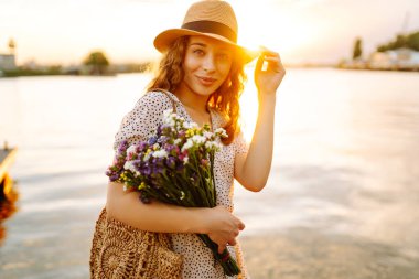 Genç bir kadın gün batımında bir buket yabani çiçekle gülüyor. Çiçek turisti bir buket çiçekle deniz manzarasının tadını çıkarıyor. Keyif ve rahatlama konsepti.