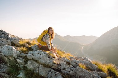 Gün batımında dağlarda sarı sırt çantalı neşeli bir kadın. Genç bayan yürüyüşçü maceranın tadını çıkarıyor ve dağ manzarasıyla özgürlüğü hissediyor. Seyahat tutkusu!