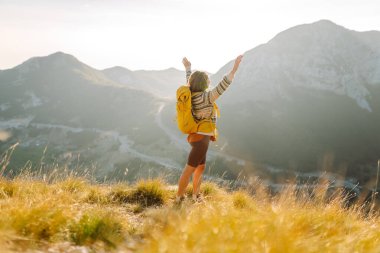 Gün batımında dağlarda sarı sırt çantalı neşeli bir kadın. Genç bayan yürüyüşçü maceranın tadını çıkarıyor ve dağ manzarasıyla özgürlüğü hissediyor. Seyahat tutkusu!