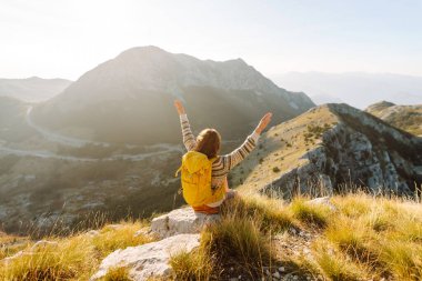 Gün batımında dağlarda sarı sırt çantalı neşeli bir kadın. Genç bayan yürüyüşçü maceranın tadını çıkarıyor ve dağ manzarasıyla özgürlüğü hissediyor. Seyahat tutkusu!