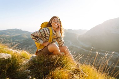 Gün batımında dağlarda sarı sırt çantalı neşeli bir kadın. Genç bayan yürüyüşçü maceranın tadını çıkarıyor ve dağ manzarasıyla özgürlüğü hissediyor. Seyahat tutkusu!