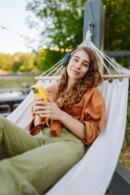 Kıvırcık saçlı genç bir kadın hamakta dinlenirken kulaklıkla müzik dinliyor. Güneşli bir parkta telefonunu kullanırken gülümseyen neşeli bir kadın. Boş zaman, blog konsepti.