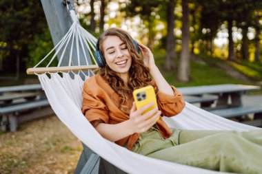 Kıvırcık saçlı genç bir kadın hamakta dinlenirken kulaklıkla müzik dinliyor. Güneşli bir parkta telefonunu kullanırken gülümseyen neşeli bir kadın. Boş zaman, blog konsepti.