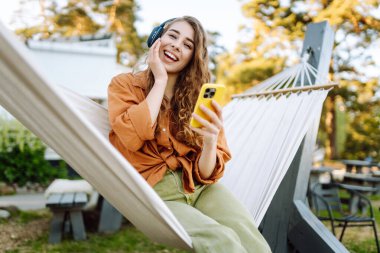 Kıvırcık saçlı genç bir kadın hamakta dinlenirken kulaklıkla müzik dinliyor. Güneşli bir parkta telefonunu kullanırken gülümseyen neşeli bir kadın. Boş zaman, blog konsepti.