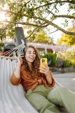 Kıvırcık saçlı genç bir kadın hamakta dinlenirken kulaklıkla müzik dinliyor. Güneşli bir parkta telefonunu kullanırken gülümseyen neşeli bir kadın. Boş zaman, blog konsepti.