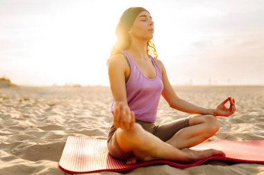 Sportif kadın gün batımı plajında yoga minderinde esneme egzersizi yapıyor. Spor kıyafetleriyle dışarıda egzersiz yapan güzel bir kadın. Spor, eğlence konsepti.