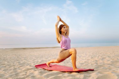 Sportif kadın gün batımı plajında yoga minderinde esneme egzersizi yapıyor. Spor kıyafetleriyle dışarıda egzersiz yapan güzel bir kadın. Spor, eğlence konsepti.