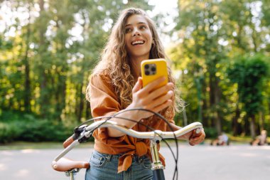 Parktaki güneşli sokakta bisiklet süren gülümseyen bir kadın. Genç bir kadın güneşli günün tadını çıkarıyor ve dışarıda eğleniyor. Hafta sonu ve günlük tutma kavramı.