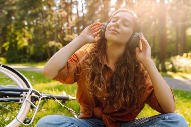 Kulaklıklı genç bir kadın parkta çimlerin üzerinde günbatımının tadını çıkarıyor. Dışarıda müzik dinleyen bisikletli ve kulaklıklı neşeli bir kadın. Etkin yaşam biçimi.