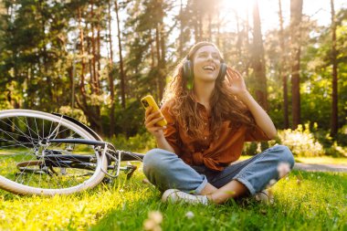 Kulaklıklı genç bir kadın parkta çimlerin üzerinde günbatımının tadını çıkarıyor. Dışarıda müzik dinleyen bisikletli ve kulaklıklı neşeli bir kadın. Etkin yaşam biçimi.