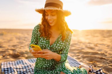 Mutlu turist, deniz manzarasında selfie çekiyor. Güzel bir kadın sahilde gün batımının tadını çıkarıyor. Telefonla, blogla. Hafta sonu, günlük tutma kavramı.
