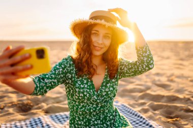 Mutlu turist, deniz manzarasında selfie çekiyor. Güzel bir kadın sahilde gün batımının tadını çıkarıyor. Telefonla, blogla. Hafta sonu, günlük tutma kavramı.