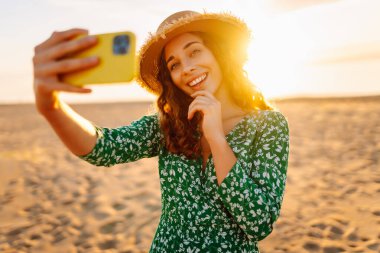 Mutlu turist, deniz manzarasında selfie çekiyor. Güzel bir kadın sahilde gün batımının tadını çıkarıyor. Telefonla, blogla. Hafta sonu, günlük tutma kavramı.