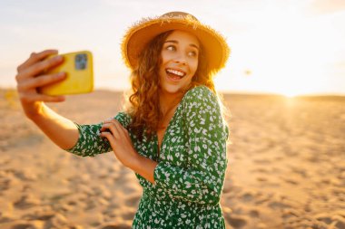 Mutlu turist, deniz manzarasında selfie çekiyor. Güzel bir kadın sahilde gün batımının tadını çıkarıyor. Telefonla, blogla. Hafta sonu, günlük tutma kavramı.